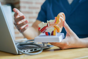 A male doctor holds a model of an eardrum while sitting at his desk. He explains a diagnosis to an unseen patient.