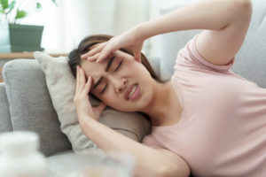 A woman in a light pink t-shirt lays on the couch wincing as she grabs her forehead with both hands and closes her eyes.