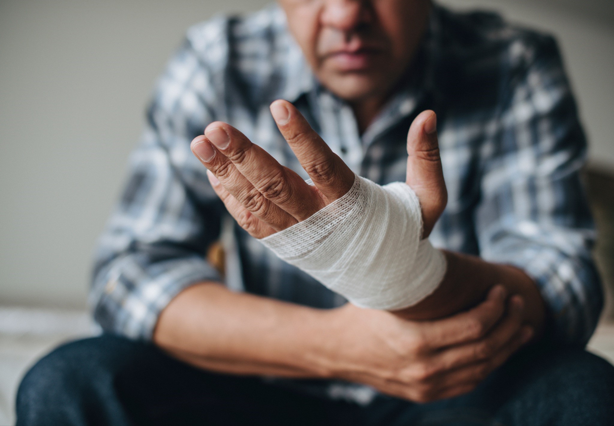 A closeup of a man sitting down and examining his hand that is wrapped in a bandage.