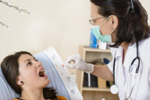 A woman at a doctor’s office gets her throat swabbed by a masked female doctor.