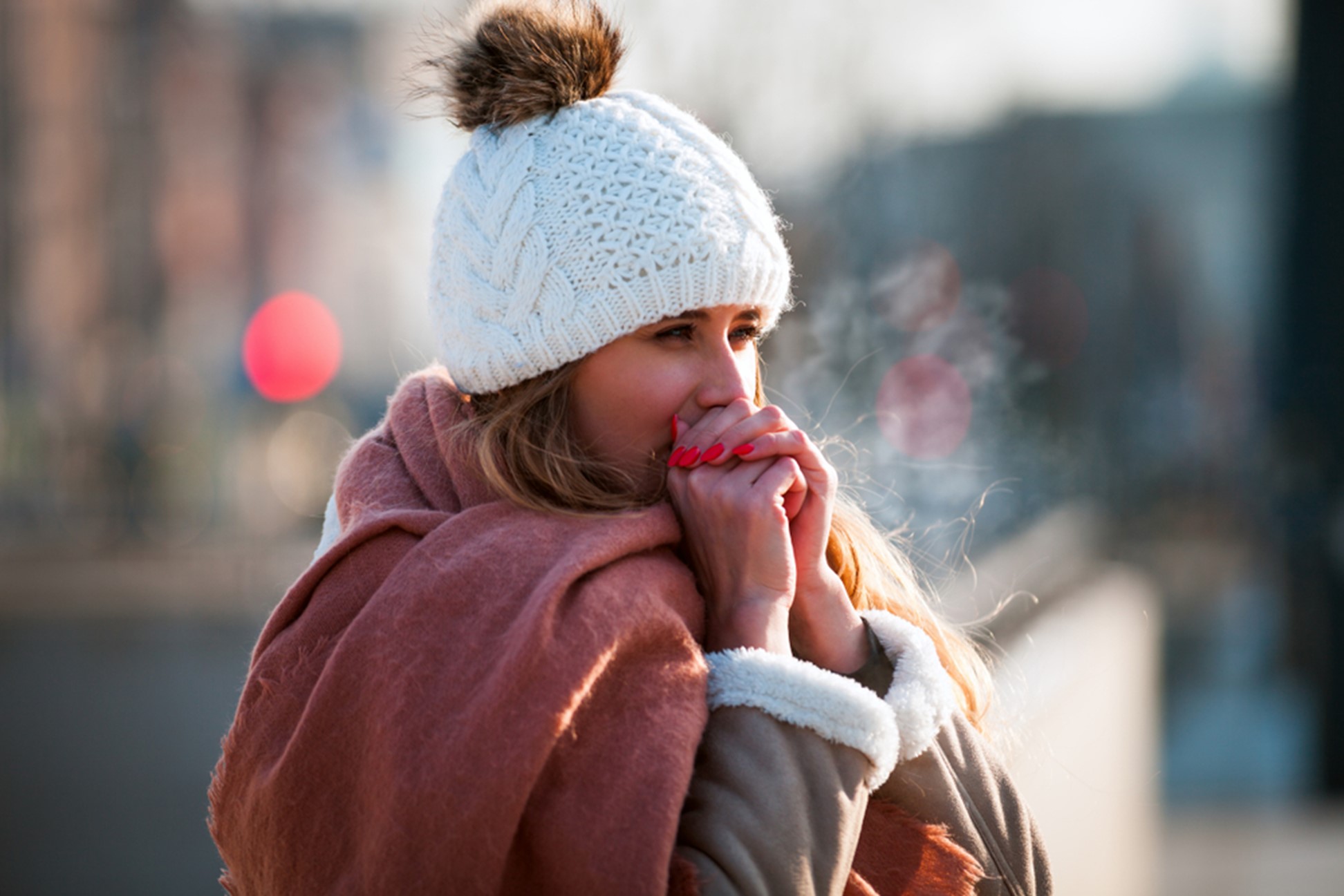 A woman in winter clothes standing outside blows into her hands to try and keep warm in the cold.