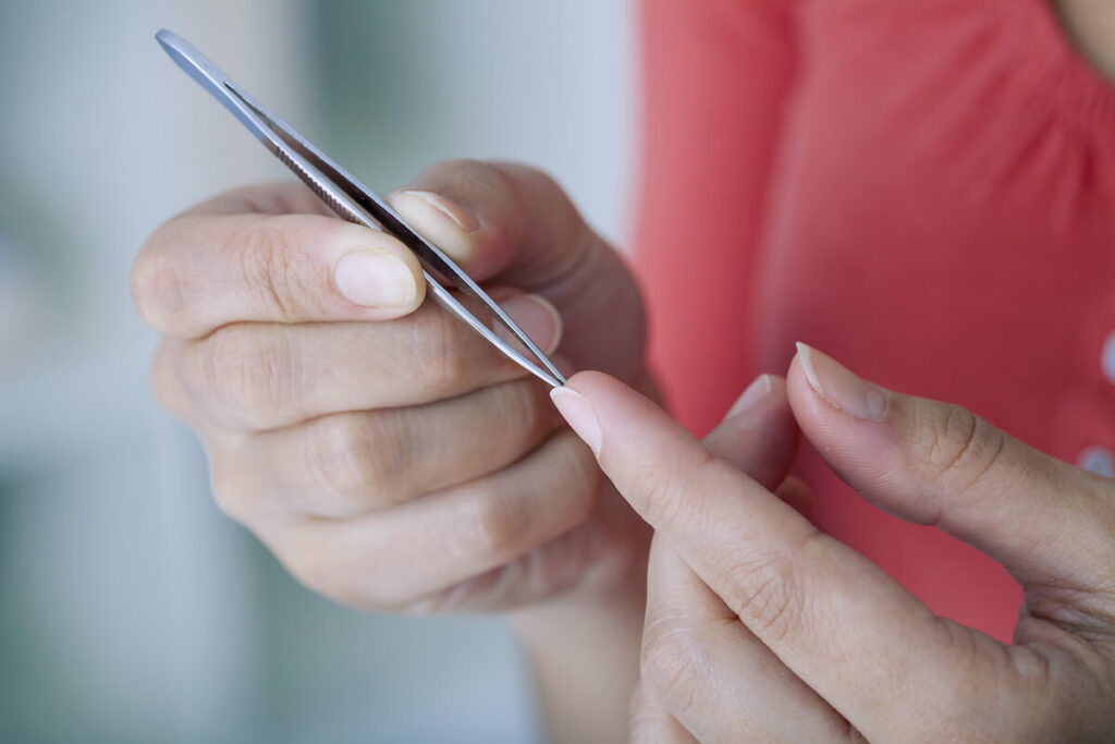 A closeup image of a woman removing a splinter from her finger with tweezers.