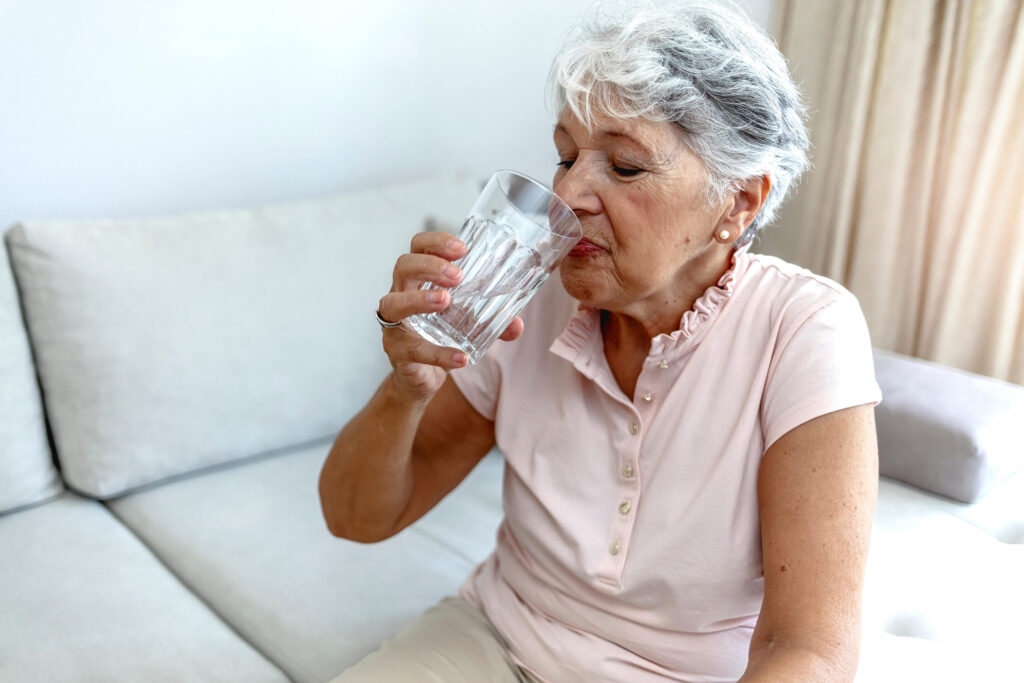 An older woman sits on the sofa drinking a glass of water.