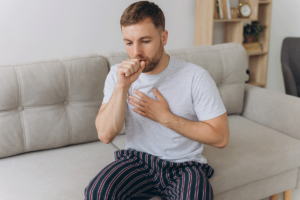 A man sits on a couch holding his chest in one hand and appears to cough into his other hand.