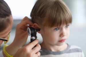 A woman wearing glasses holds an otoscope and looks inside a girl’s right ear.