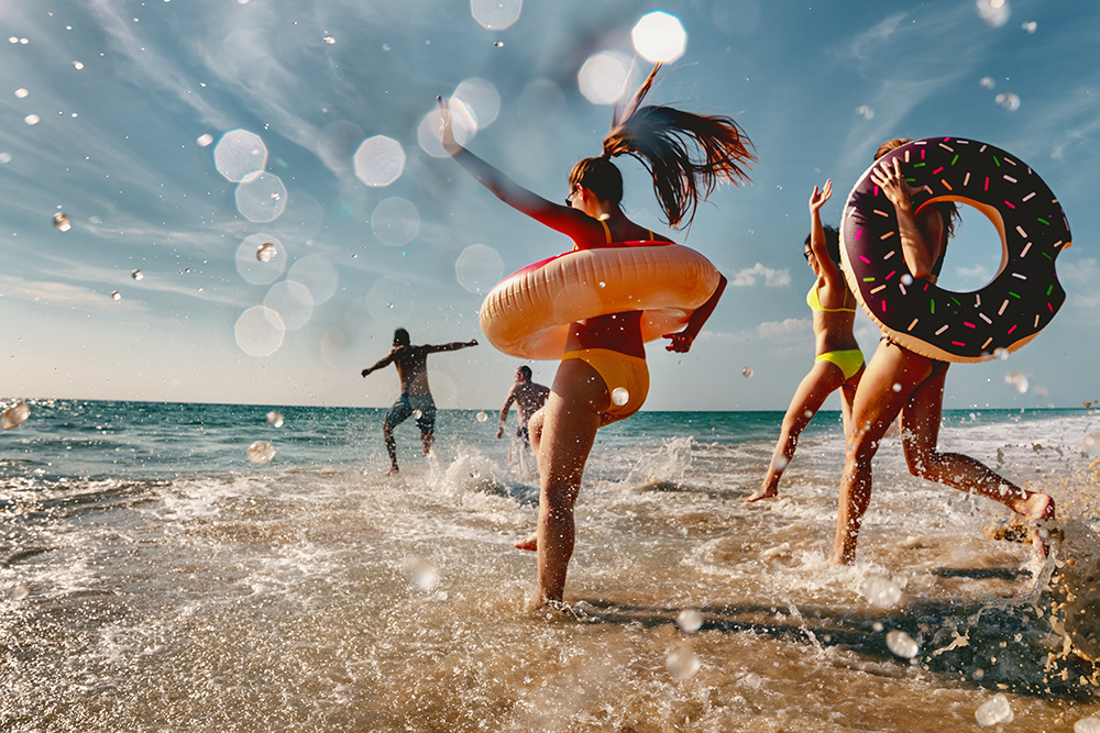 A group of young adults running into the ocean with inflatable swim rings on a sunny beach day, splashing through the water.