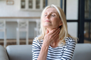 An older woman sits on the sofa with her eyes closed and a hand on her throat.