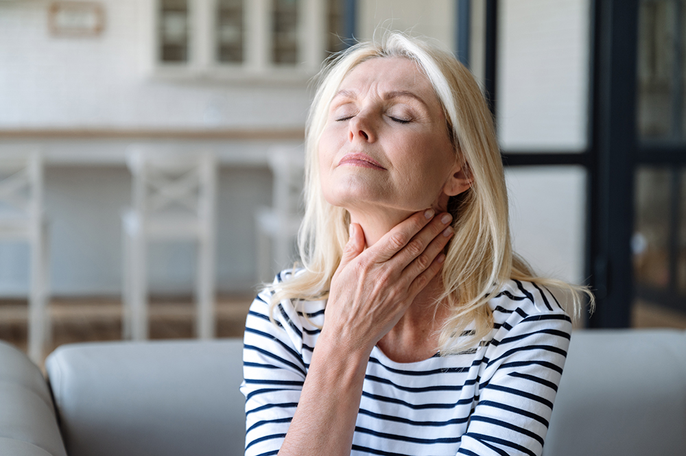 An older woman sits on the sofa with her eyes closed and a hand on her throat.